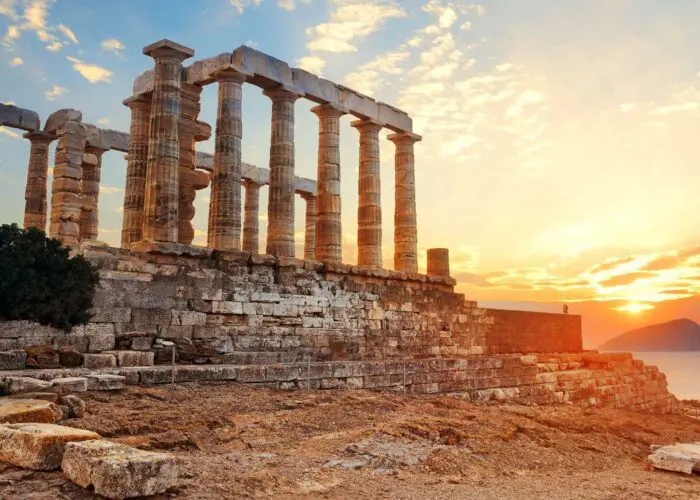The ancient stone columns of the Temple of Poseidon at Cape Sounion, Greece, stand against a sunset sky with the sea and a distant island in the background—one of the must-see places to visit in Greece.