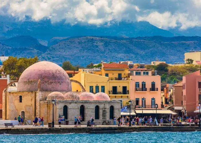 A historic stone mosque with a pink dome stands by the waterfront, surrounded by colorful buildings and a crowd of people—an iconic scene to experience on your Greece tour, with mountains and clouds in the background.