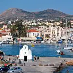 Aegina Island, Greece - Charming small white chapel on a waterfront pier in Aegina Island, with people strolling nearby, sailboats docked in the marina, and a hillside town beyond—a perfect Athens island day trip spot for your 2026 guide.
