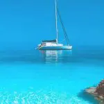 A white sailboat floats on calm, clear turquoise water near rocky shorelines under a bright blue sky, offering a glimpse of the serene beauty you can experience on Paros Boat Tours.