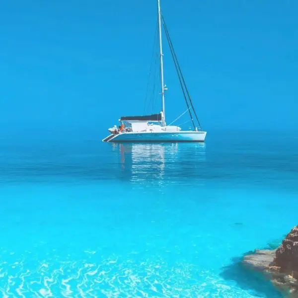 A white sailboat floats on calm, clear turquoise water near rocky shorelines under a bright blue sky, offering a glimpse of the serene beauty you can experience on Paros Boat Tours.