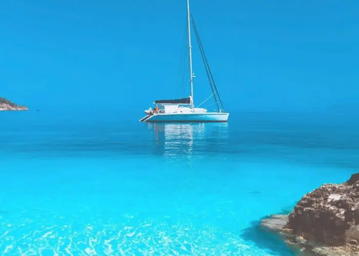 A white sailboat floats on calm, clear turquoise water near rocky shorelines under a bright blue sky, offering a glimpse of the serene beauty you can experience on Paros Boat Tours.