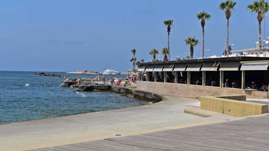 People enjoy a sunny day at Paphos Harbour's waterfront promenade, where palm trees sway and a yacht glides by in the distance, making it one of the top attractions and things to do.