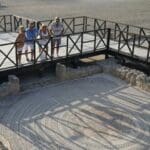 People stand on a wooden platform in Western Cyprus, overlooking an ancient mosaic nestled among stone ruins. This site, rich with living traditions, offers a glimpse into the vibrant history that shaped this land.
