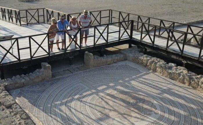 People stand on a wooden platform in Western Cyprus, overlooking an ancient mosaic nestled among stone ruins. This site, rich with living traditions, offers a glimpse into the vibrant history that shaped this land.