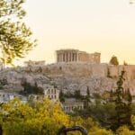 View of the Acropolis in Athens, Greece, at sunset. The Parthenon is visible at the top of the hill, surrounded by trees and ancient ruins.