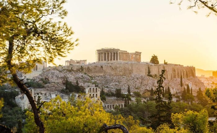 View of the Acropolis in Athens, Greece, at sunset. The Parthenon is visible at the top of the hill, surrounded by trees and ancient ruins.