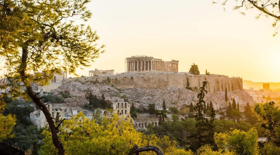 View of the Acropolis in Athens, Greece, at sunset. The Parthenon is visible at the top of the hill, surrounded by trees and ancient ruins.