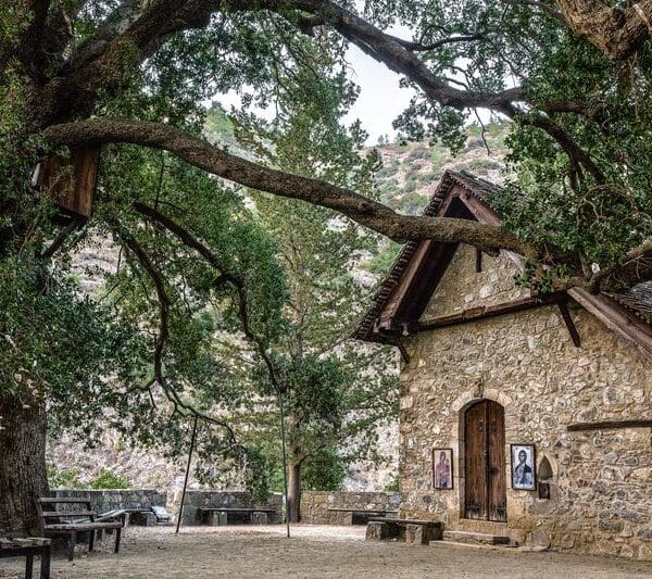 A small stone building with wooden doors and windows stands under large, old trees with a bench and framed pictures nearby, against a backdrop of hills and greenery.