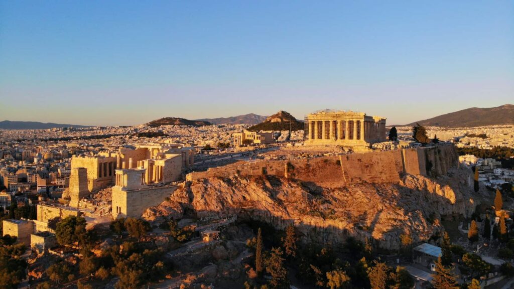 Aerial view of the Acropolis in Athens, Greece, featuring the Parthenon and other ancient structures, set against a backdrop of the city and distant mountains at sunset.