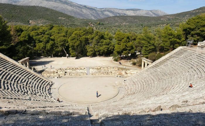 Ancient amphitheater with tiered stone seating and a central stage, surrounded by forested hills and mountains. Sparse visitors are scattered around the seating area and stage.