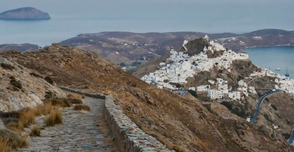Rocky path leading to a hillside village with white buildings on Serifos Island, surrounded by the sea and barren landscape.