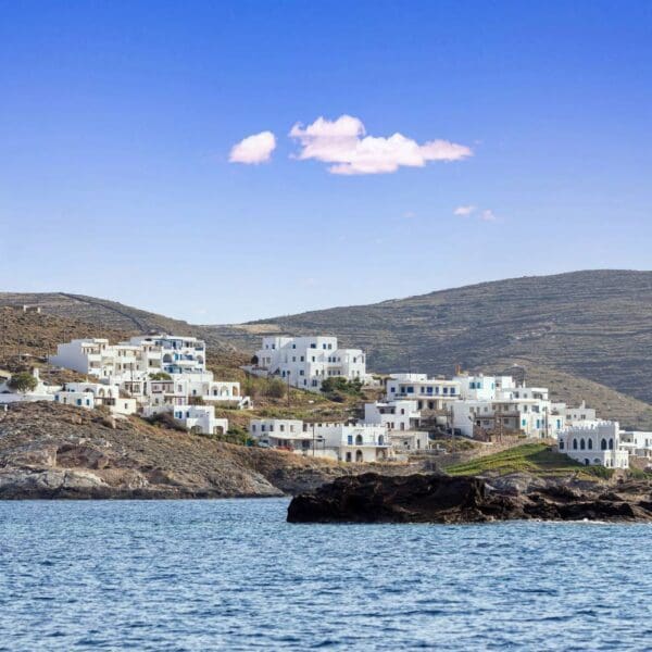 White houses dot a hillside by the sea under a clear blue sky on picturesque Kythnos Island, with rugged terrain in the background.