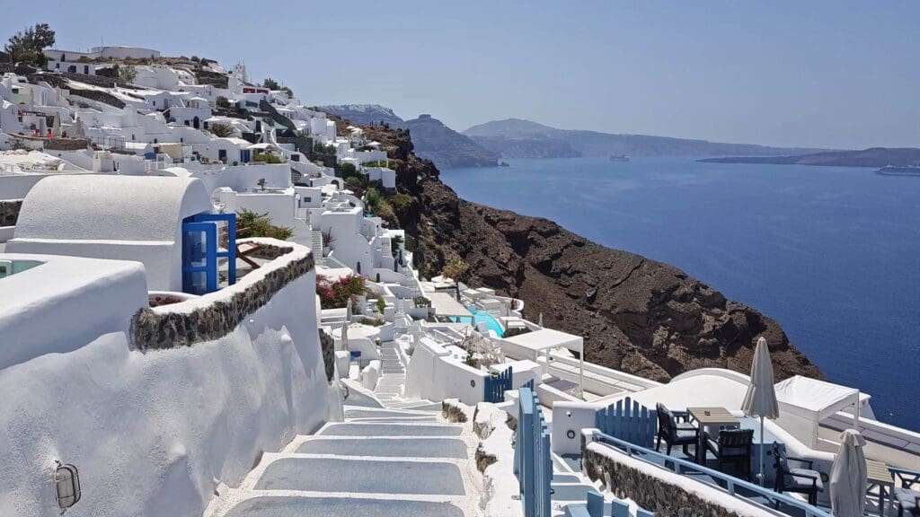 White buildings on a hillside overlook the blue sea, with a clear sky above and distant hills in the background, capturing the essence of Santorini Island. 