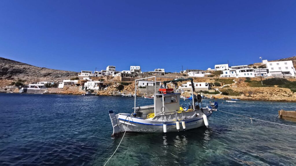 A small fishing boat is docked in the clear blue waters of Sifnos Island, with white buildings scattered on a rocky hillside under a bright blue sky, inviting exploration.