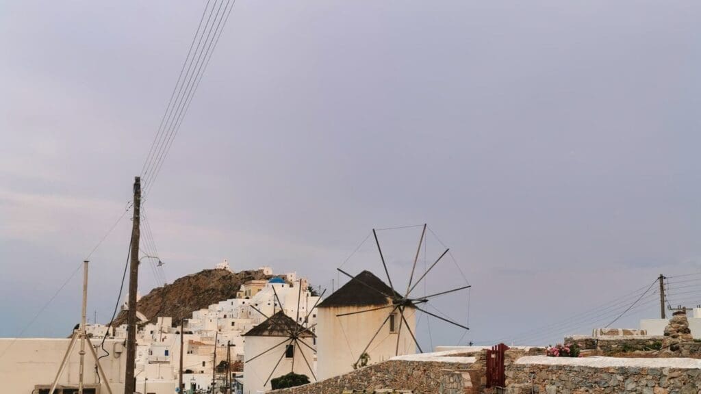 White buildings and a traditional windmill on a hill under a cloudy sky evoke the charm of Serifos Island, with telephone wires and stone walls in the foreground. This scenic view invites exploration, blending perfectly into any travel guide's picturesque narrative.