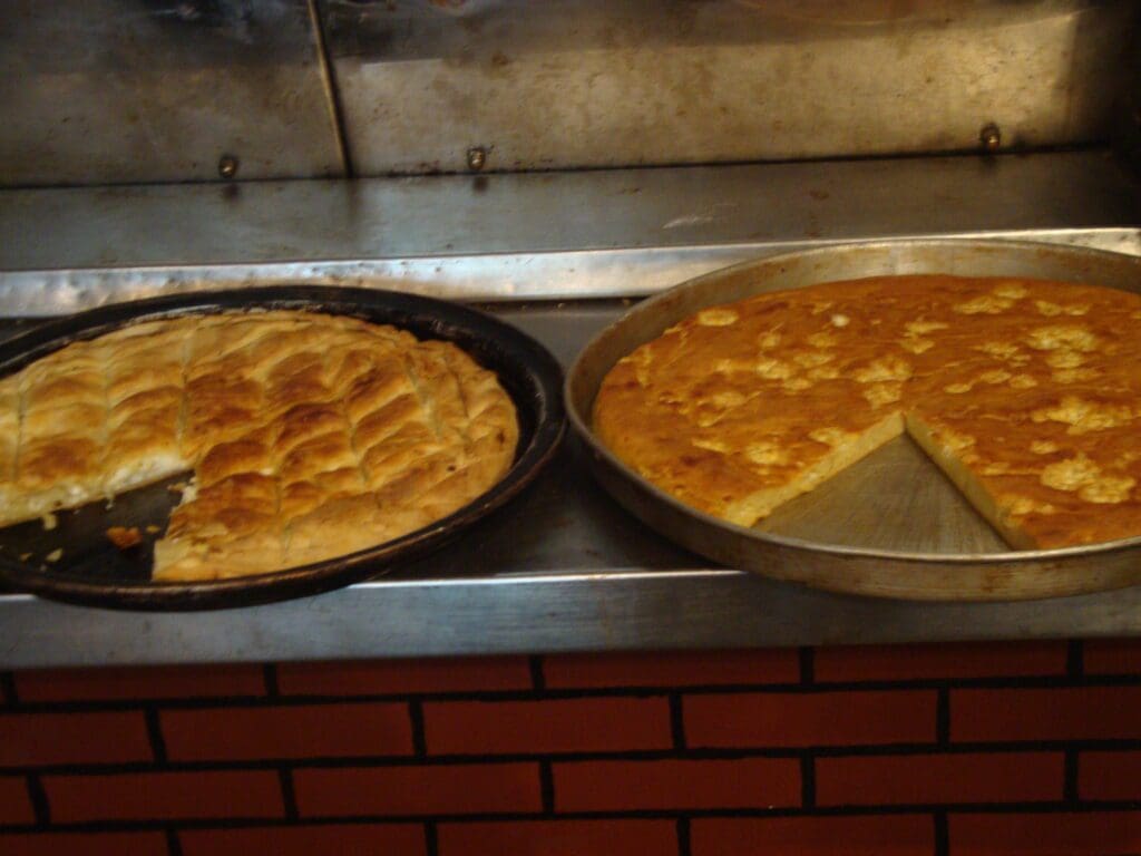 Two round baking trays rest on the counter, one holding a cut puff pastry pie reminiscent of Greek street food delights, while the other tray displays a perfectly sliced flatbread.