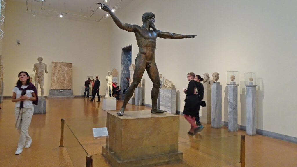 In a room frequented by visitors pondering their next destination, a museum exhibit boasts a grand bronze statue surrounded by other stone sculptures. It's the perfect stop after exploring how many days in Athens you wish to enjoy.