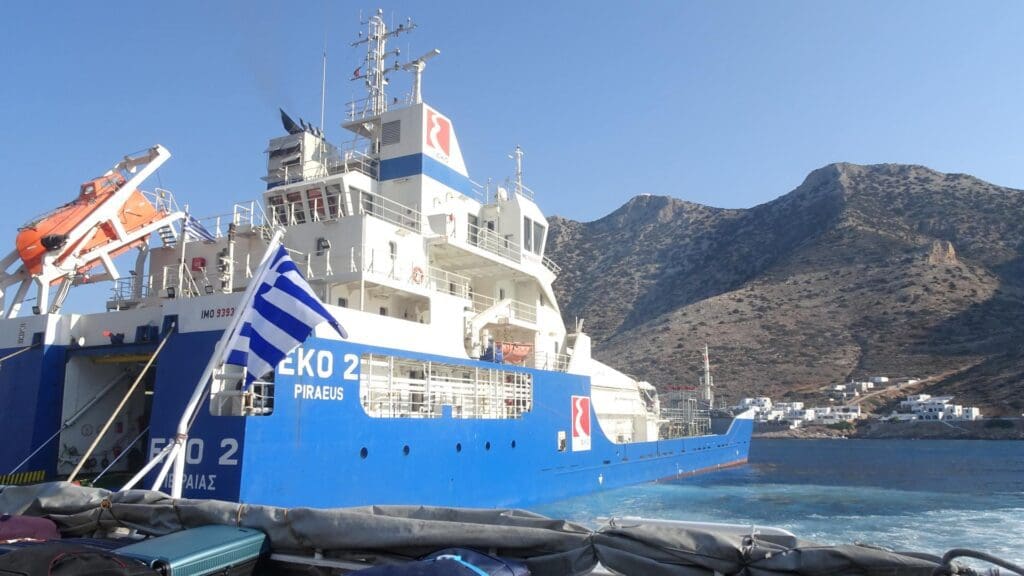A large blue and white ferry named "Naxos 2" is docked by a mountainous shoreline, with a Greek flag visible, ready for itineraries perfect for the adventurous Greek island-hopping traveler.