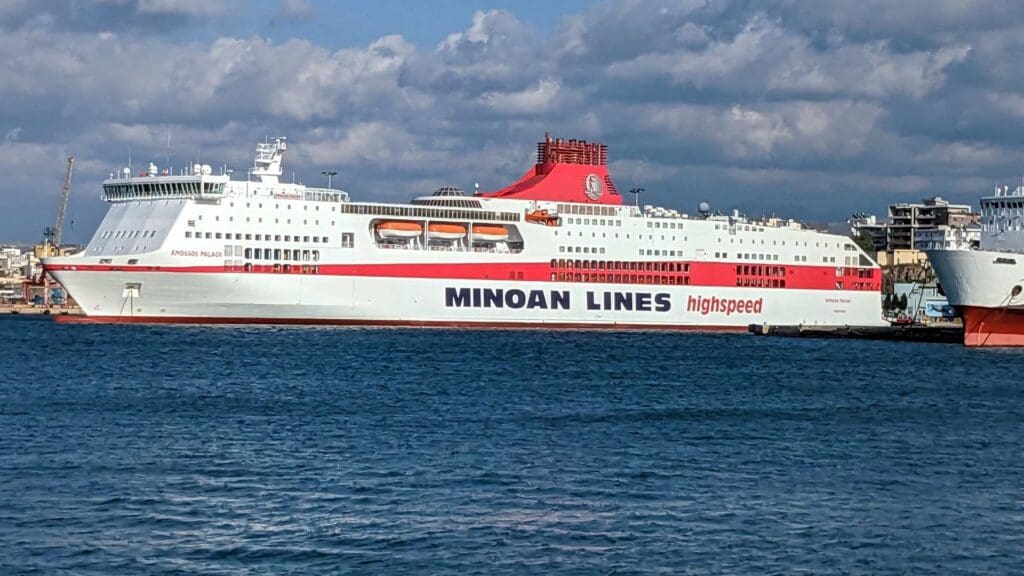 A large ferry named "Minoan Lines" is docked at a port under a partly cloudy sky, ready to whisk travelers away to the sun-kissed Heraklion beaches.