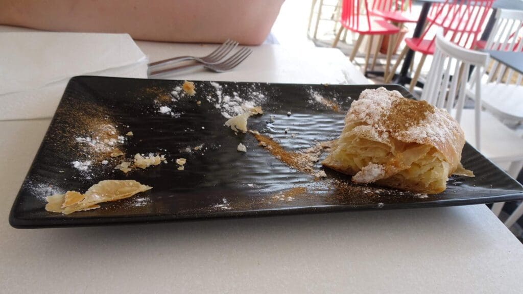 A mostly eaten pastry, reminiscent of Greek street food, dusted with powdered sugar and cinnamon on a black rectangular plate with scattered crumbs, rests on a white table. A fork and napkins linger in the background.