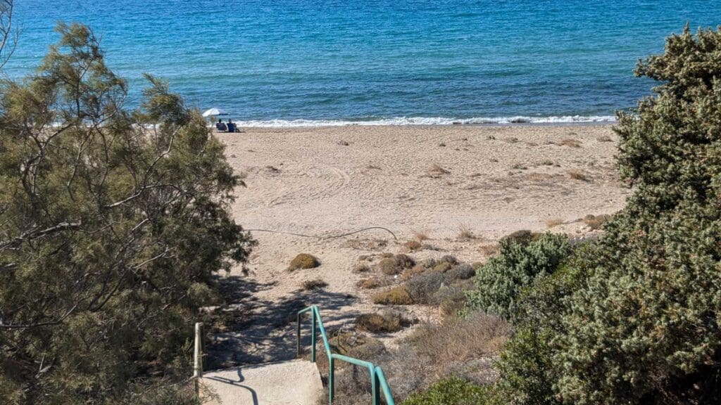 Discover a serene pathway leading to a sandy beach, where two people relax under a white umbrella near the shoreline. Surrounded by lush vegetation, this hidden gem is one of Milos Beaches' must-see spots for those seeking tranquility and natural beauty.