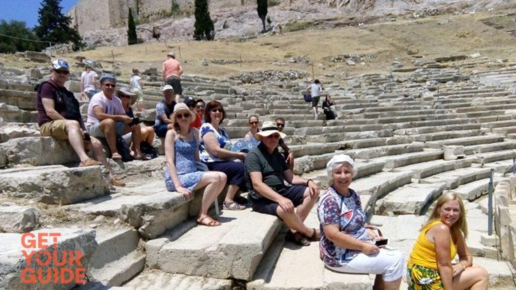 A group of people sitting on ancient stone steps, surrounded by rocky terrain and trees, possibly visiting the Acropolis.