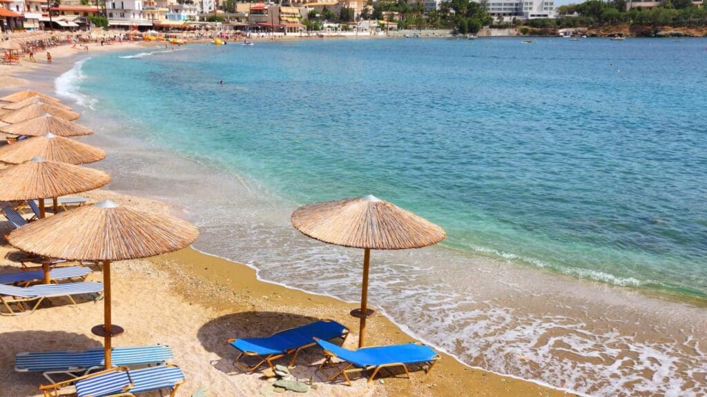 Sandy beach with blue lounge chairs and straw umbrellas, overlooking the clear blue waters of Heraklion beaches and a distant shoreline with buildings.