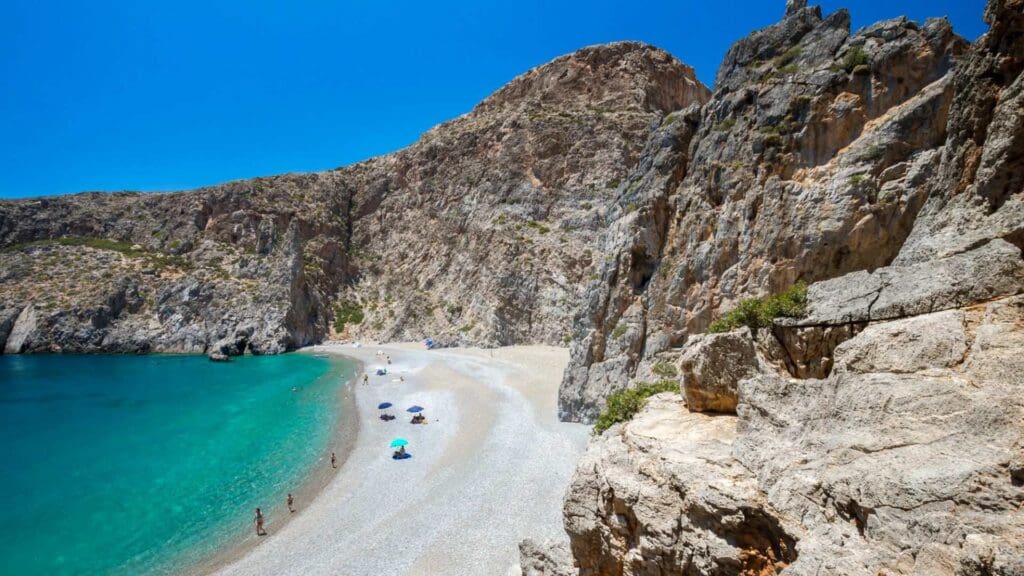 A rocky beach near Heraklion with turquoise water, towering cliffs, and a few people sunbathing under colorful umbrellas.