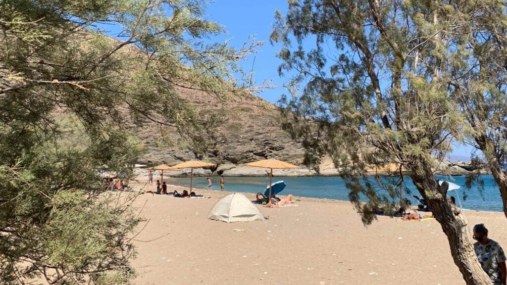 A sandy beach on Sikinos Island features scattered trees, a tent, and people relaxing under umbrellas. Hills and the blue sea stretch into the background beneath a clear sky.