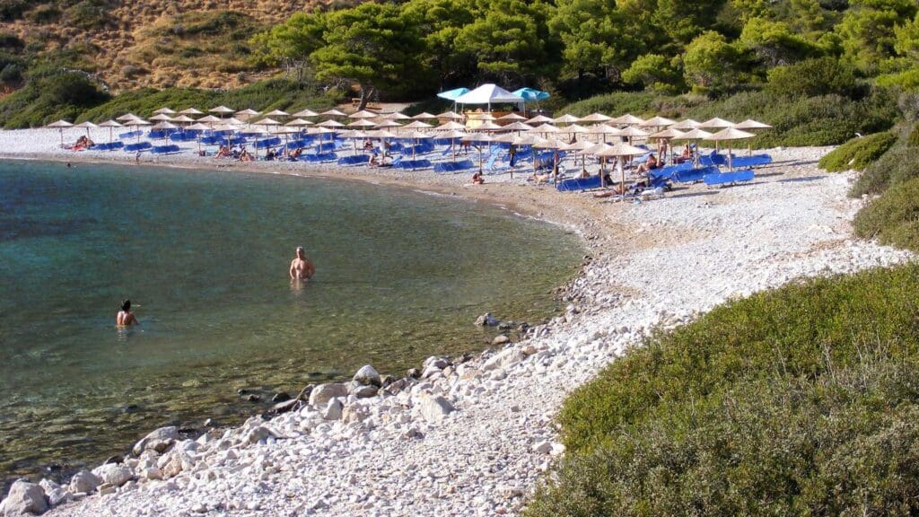 Nestled among the lush greenery, Hydra beaches offer a stunning pebble beach adorned with sun loungers and umbrellas. Visitors can be seen swimming in the crystal-clear waters and lounging under the warm sun.