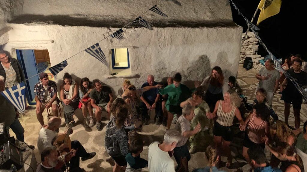 A group of people dance and sit outside a white stone building adorned with Greek flags on Sikinos Island at night.