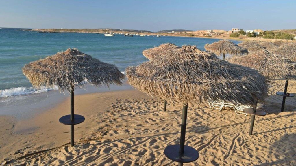 A sandy beach with straw umbrellas, clear blue water, and a distant view of boats and buildings on the horizon captures the charm of Naoussa, Paros.