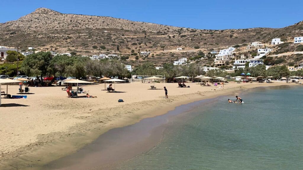 On a sunny day, the sandy beach of Sikinos Island offers clear waters, surrounded by hills and scattered houses. People relax under umbrellas and swim in the sea's inviting embrace.