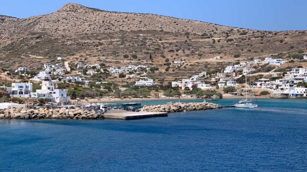 A coastal village on Sikinos Island features white buildings perched on a rocky hillside, overlooking a small dock and a sailboat gliding across the blue waters under a clear sky.