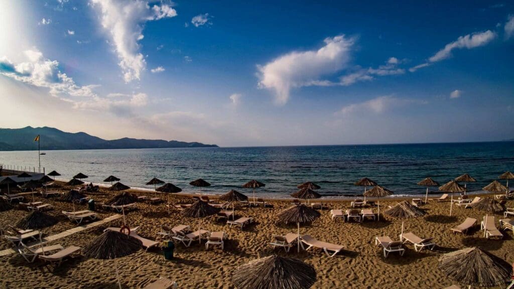 Sandy beach with rows of empty sun loungers and straw umbrellas facing the calm sea under a blue sky with few clouds, reminiscent of Heraklion beaches. Hills are visible in the distance.