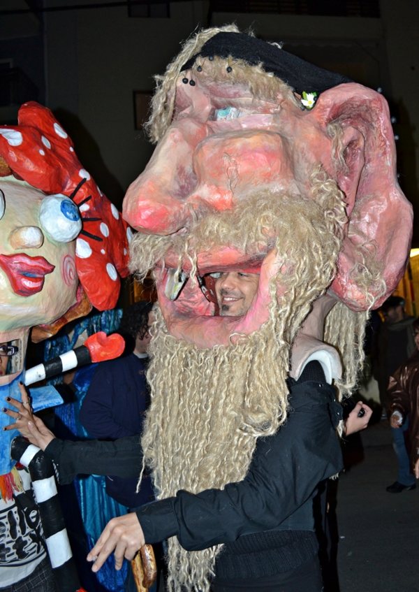 During the lively Apokries celebration, a person wearing a large, whimsical mask with exaggerated features and long hair stands beside another in a vibrant costume.