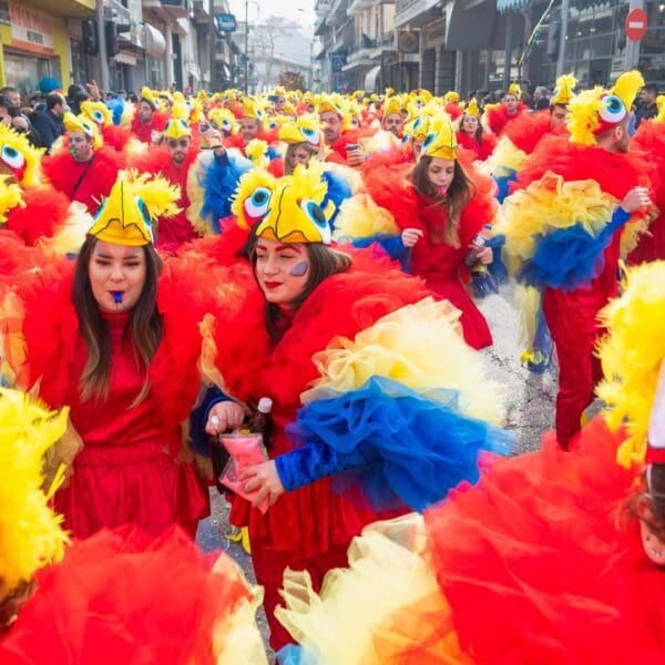 A large group of people dressed in vibrant bird costumes with colorful feathers parade down a busy street, capturing the festive spirit of Apokries.