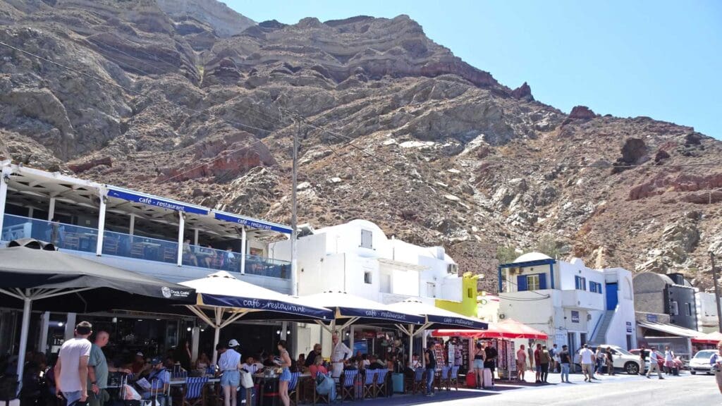 People sit at outdoor cafes with umbrellas near rocky cliffs under a clear blue sky, enjoying the views not far from the Santorini ferry port.