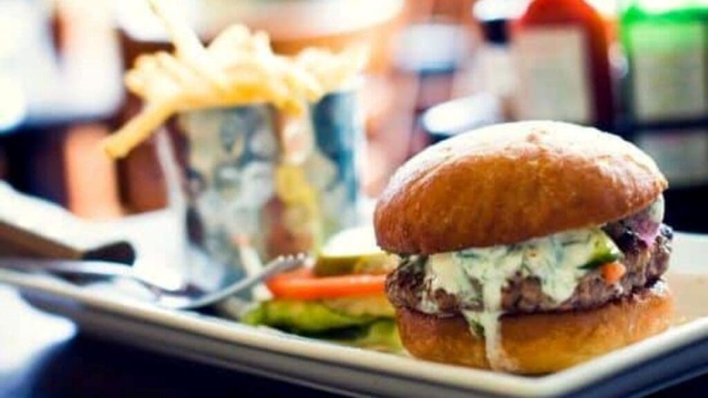 A burger with creamy sauce on a bun sits on a plate, echoing the flavors of Greek street food, with a side of sliced vegetables. Fries are served in a cup nearby, completing this delightful culinary experience.