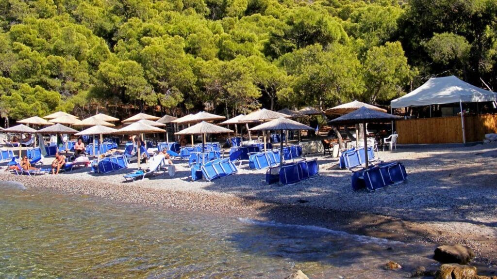 A serene beach scene on one of Hydra's beaches features empty blue lounge chairs and straw umbrellas dotting the pebbly shore, embraced by lush green trees. A few people stroll nearby, with a tent structure standing in the background.