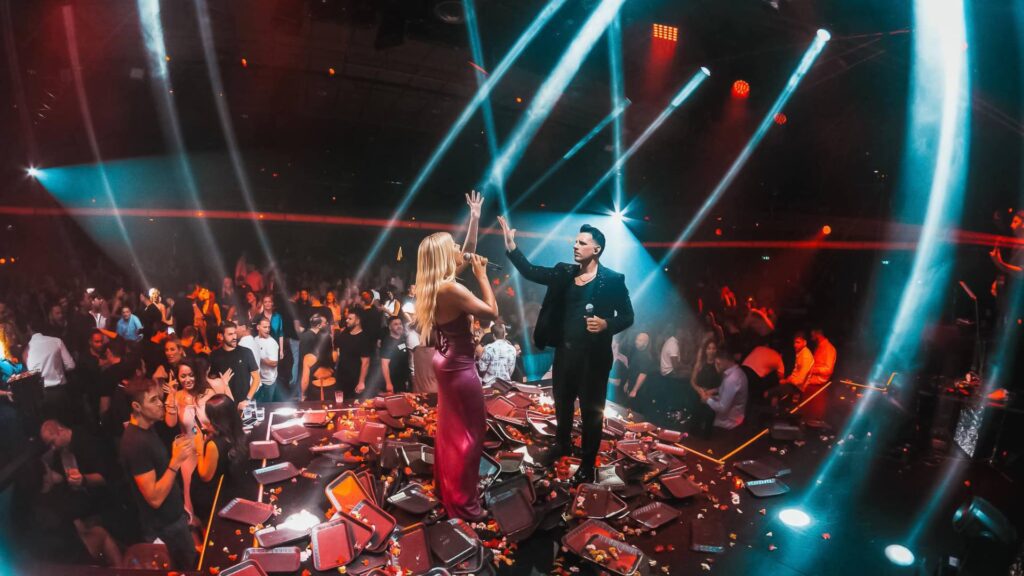 In Athens, two performers on stage high-five during a vibrant Christmas concert, surrounded by an energetic crowd and colorful lights—truly one of the best things to do this festive season.