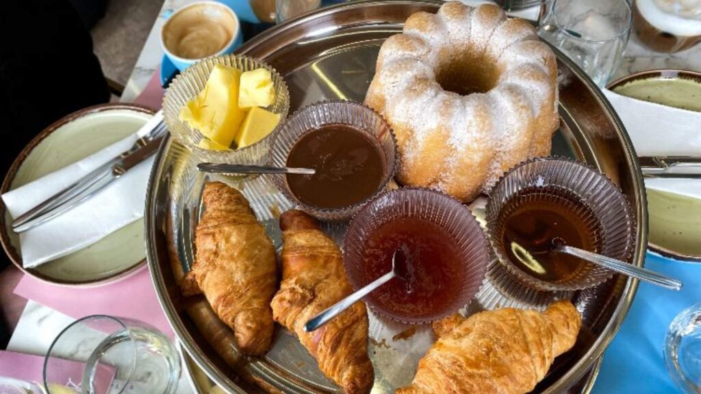 A plate with croissants, a powdered bundt cake, butter slices, chocolate, jam, and honey awaits beside napkin-wrapped cutlery and coffee—a delightful start before exploring the best things to do this Athens Christmas.
