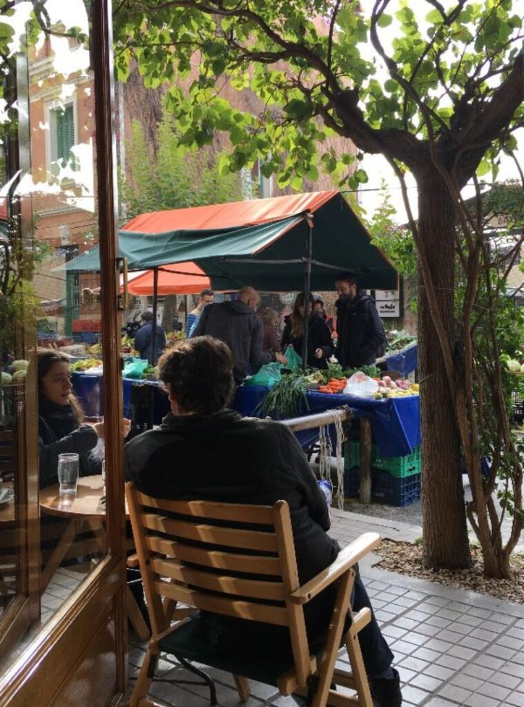 People browse and sell fresh produce at an outdoor market stall under a canopy. Tables display various fruits and vegetables near a cafe with outdoor seating, offering the perfect spot to enjoy seasonal delights during Christmas in Athens.