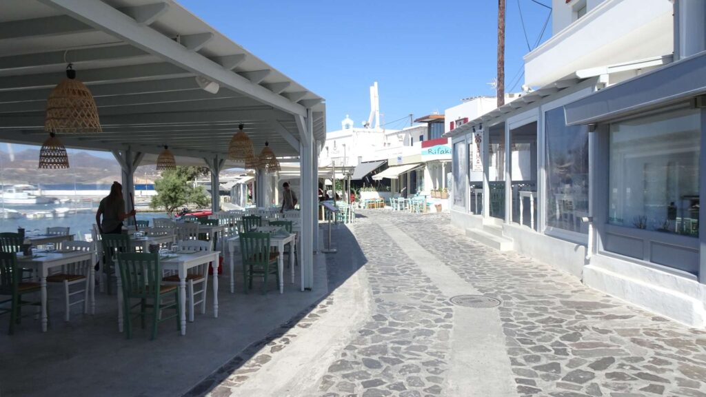 A cobblestone street on Paros is lined with outdoor cafes and shops under a clear blue sky. Some tables and chairs are arranged in a covered area, inviting visitors to relish the island's charm.