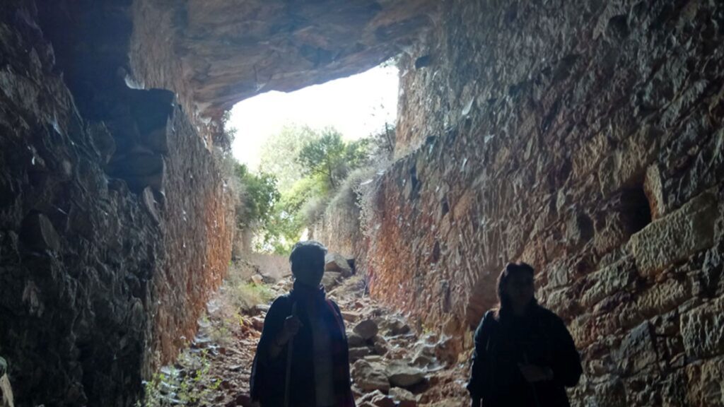Two people stand inside a rock tunnel with rough, stone walls. The sunlight and greenery of Naoussa, Paros are visible through the opening at the far end.