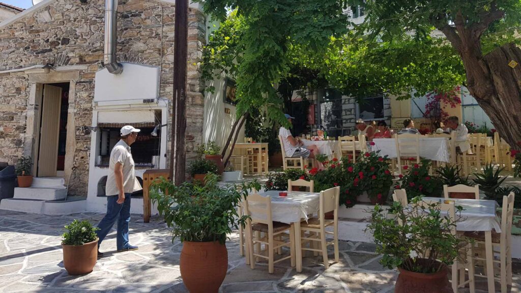 In a charming outdoor restaurant on Naxos, wooden tables and chairs nestle under lush green foliage. A person strolls by as a group enjoys their meal near stone walls and vibrant potted plants, capturing the island’s serene ambiance.