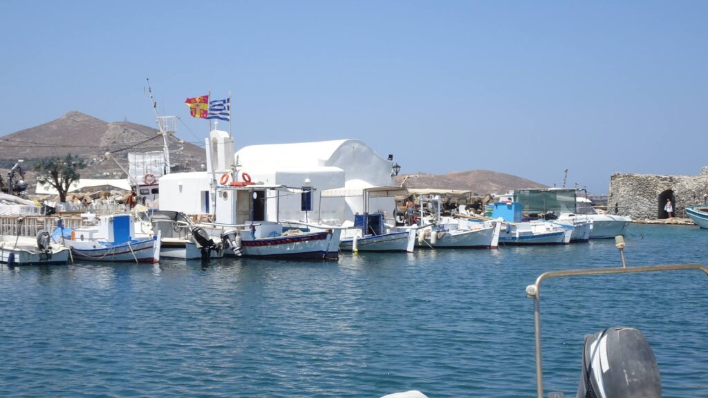 Nestled in the charming village of Naoussa, Paros, a small harbor hosts several fishing boats moored beside a white building, framed by a mountainous landscape under a clear blue sky. Vibrant flags flutter proudly atop the building.