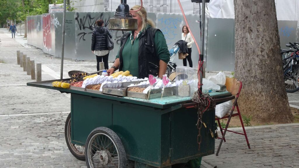 A street vendor in Athens at Christmas sells various snacks and sweets from a green cart on a cobblestone sidewalk, offering visitors delightful treats to enjoy as they explore the city's festive attractions.