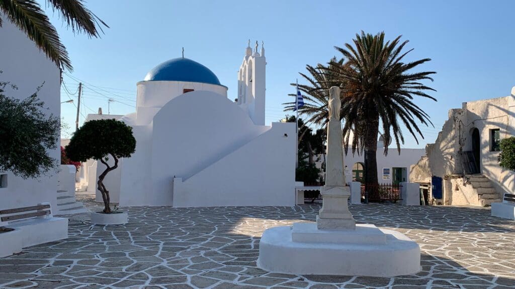 A white church with a blue dome graces a sunlit courtyard on Sikinos Island, surrounded by palm trees, with a flag and statue in the foreground.